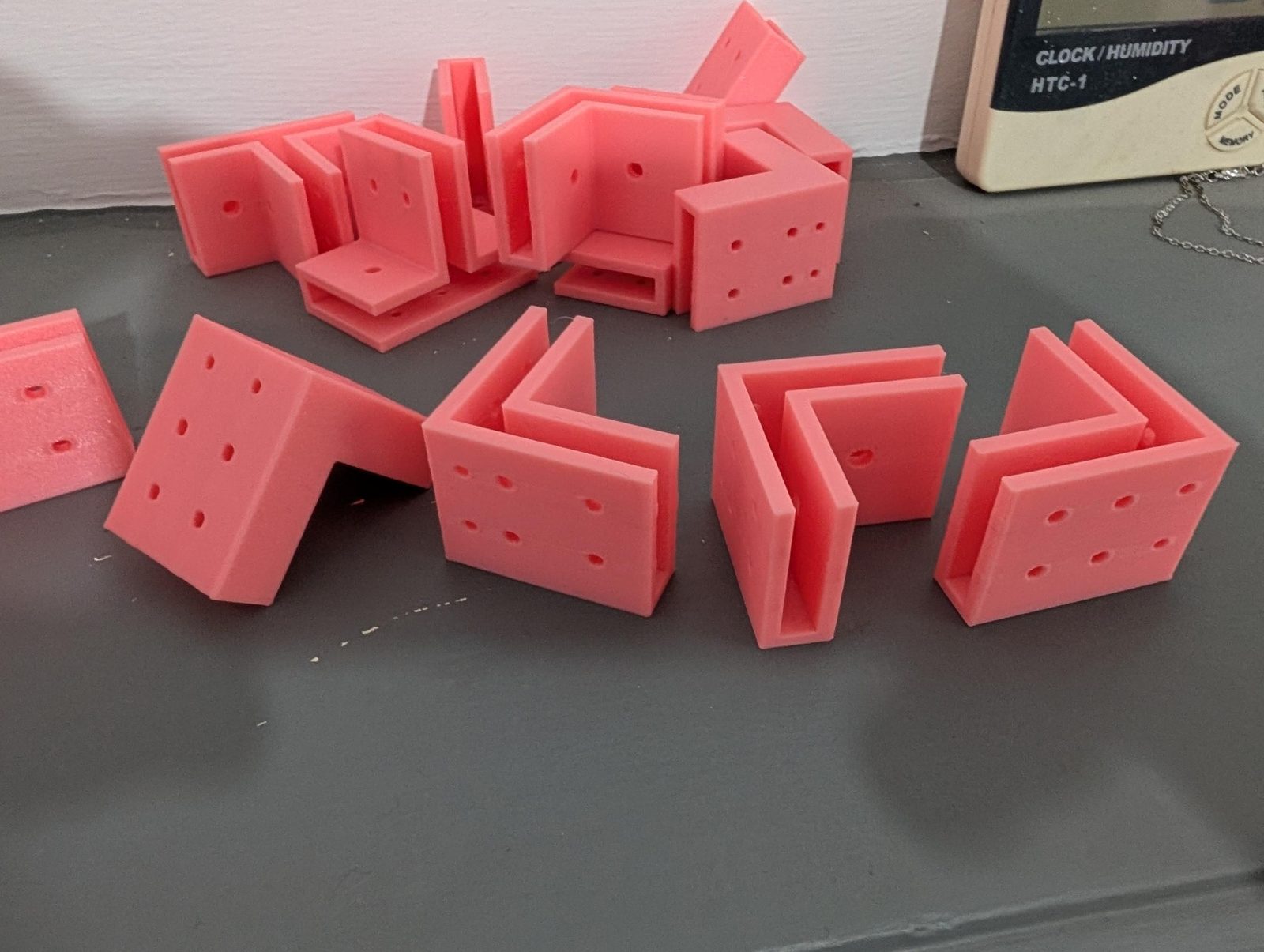 A row of pink printed connectors: a T-joint lying on its back wall showing six holes, and three L-corners with nail and screw holes in their outer faces.