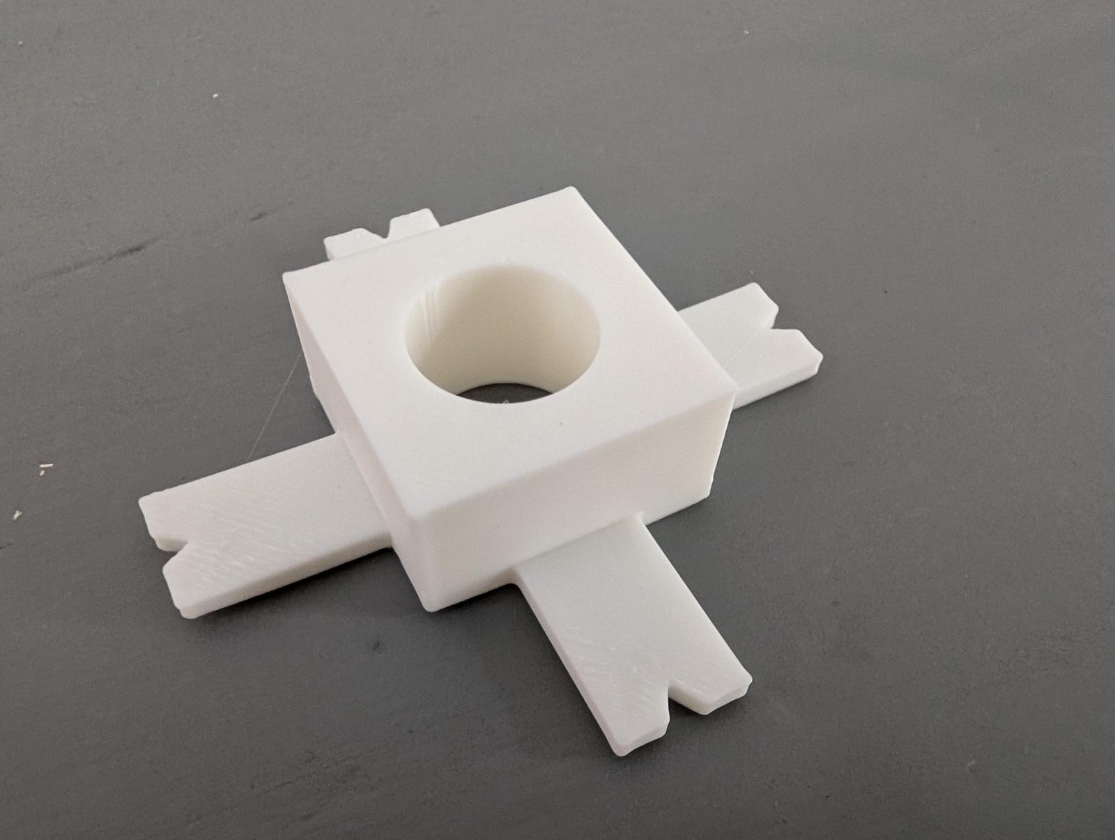 The printed drill guide on a grey surface: a white square block with a round hole through the middle and four flat feet with V-notches at their tips.
