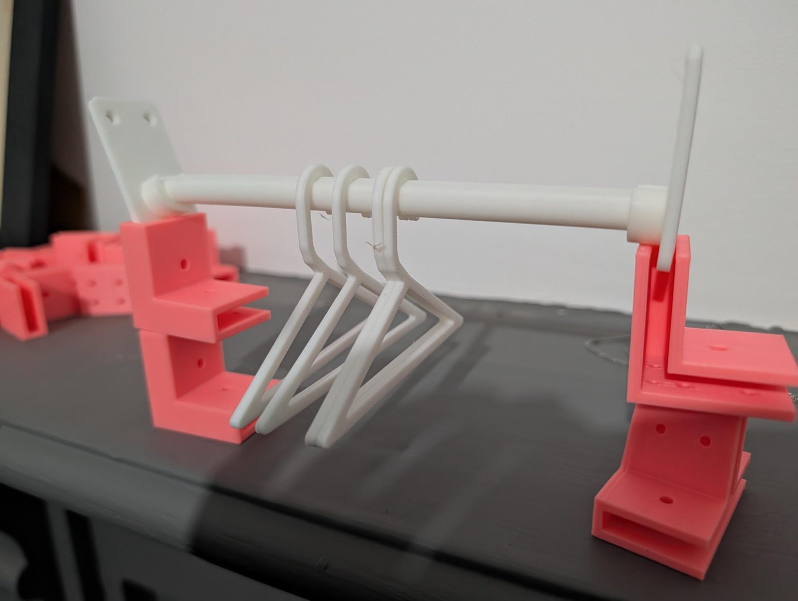 A white printed rail resting in two white brackets, with three white coat hangers hanging from it. The brackets stand on pink printed board connectors.